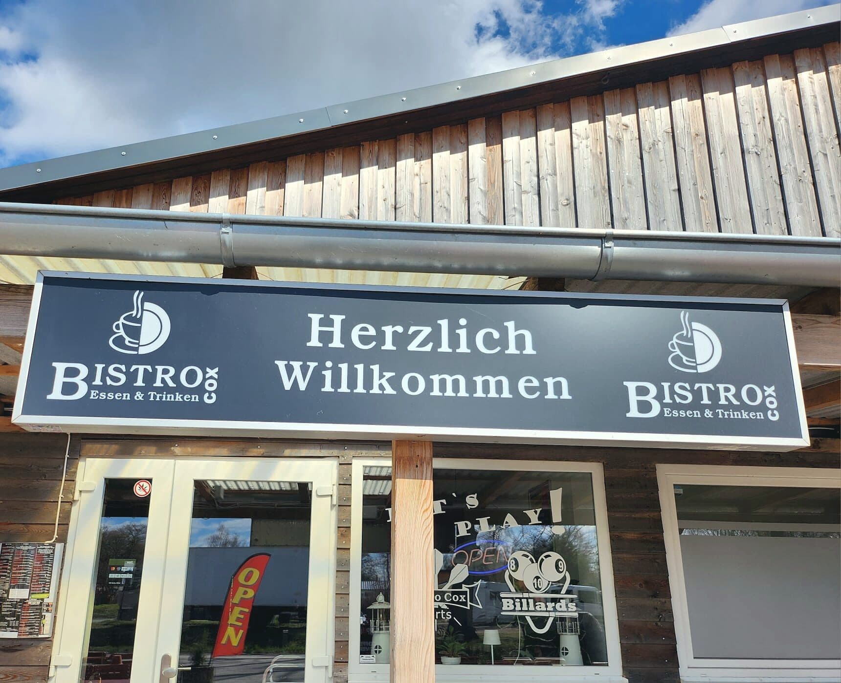 Außenansicht des Cox Bistro: Holzfassade, Leuchtschild «Herzlich Willkommen» mit Logo, Eingangstür und Fenster mit Billard-Schriftzug