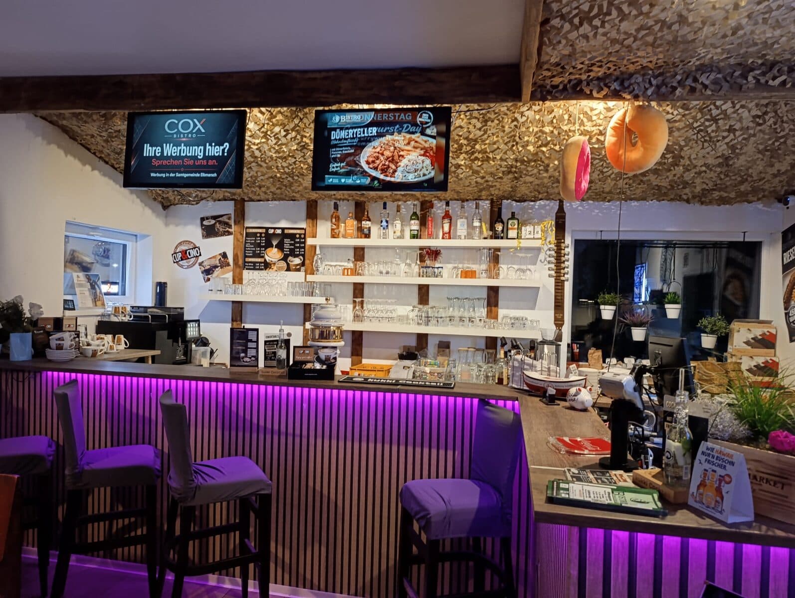 Theke mit lila Beleuchtung, Barhockern und Gläserregal, darüber zwei Bildschirme